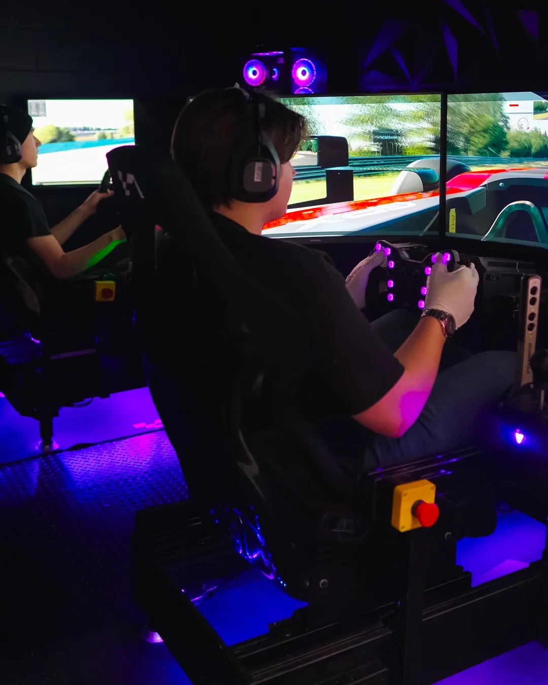 Driver in a Simul8Racing cockpit with triple monitors and purple lighting.